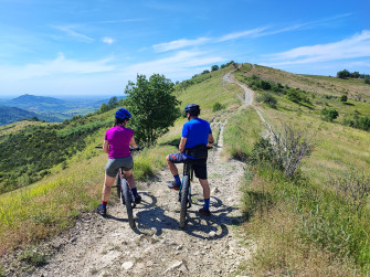 Due ciclisti in sella alle loro e-bike ammirano il paesaggio a 360° da una collina, affacciati sull'Oltrepò Pavese